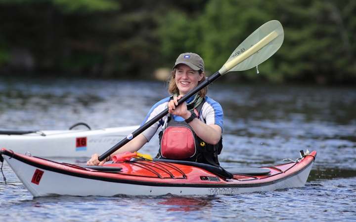 A person smiles as they paddle a kayak on calm water. 